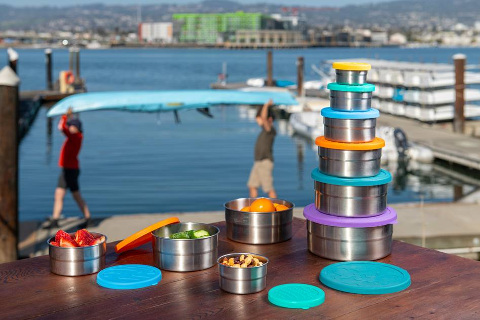 Bamboo and stainless steel reusable snack containers arranged on a wooden kitchen table with fresh fruits